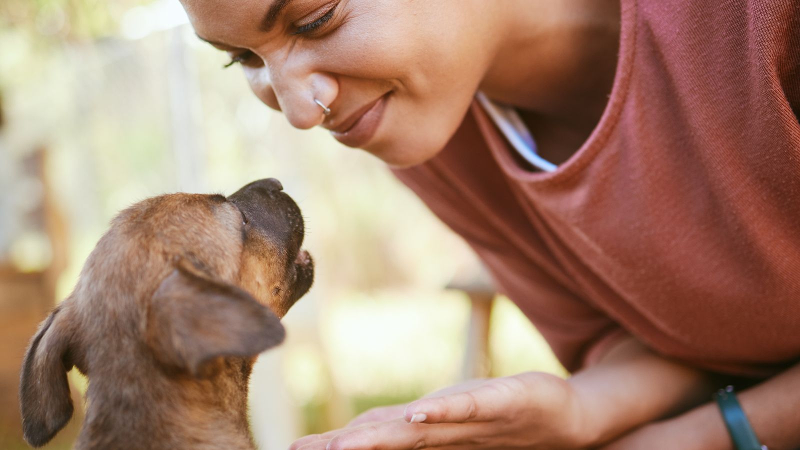 A smiling woman leaning down nose to nose with a small brown puppy in a garden